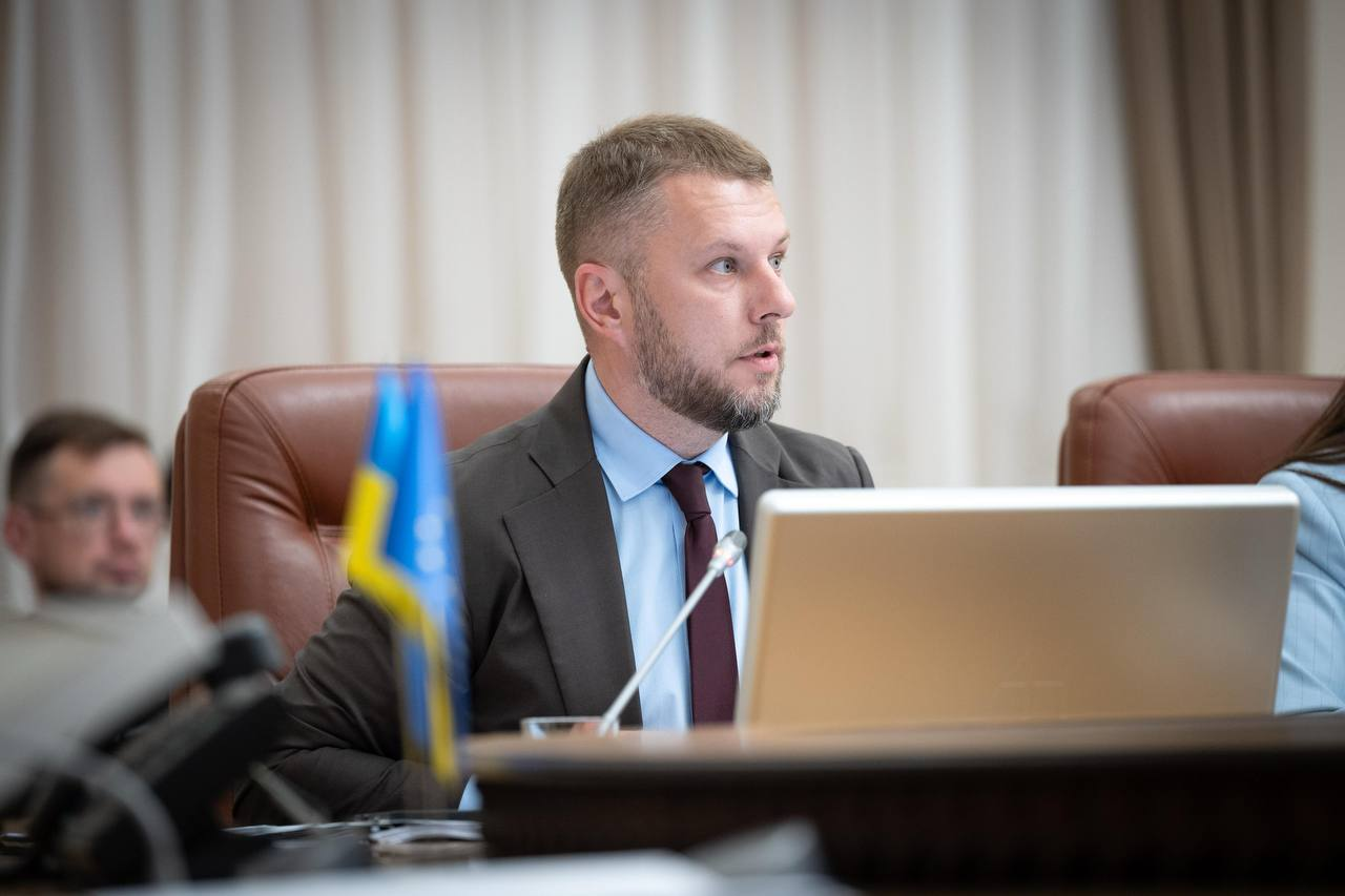 Чоловік у діловому костюмі, блакитній сорочці та краватці сидить у кріслі за столом засідань поруч із відкритим ноутбуком і настільним мікрофоном. На передньому плані видно невеликий прапорець України, а на розмитому задньому плані — іншого учасника зустрічі.