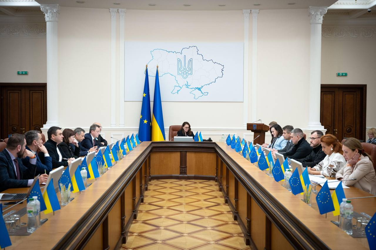 Група людей у діловому одязі сидить за великим П-подібним столом під час офіційного засідання. Столи прикрашені прапорцями України та ЄС, а на центральній стіні розміщено контурну карту України з Тризубом.
