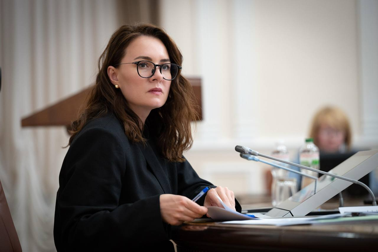 Жінка в окулярах та чорному піджаку сидит за круглим столом засідань, тримаючи в руках документи та ручку. Перед нею встановлені настільні мікрофони, а на розмитому задньому плані видно іншу учасницю заходу та трибуну.