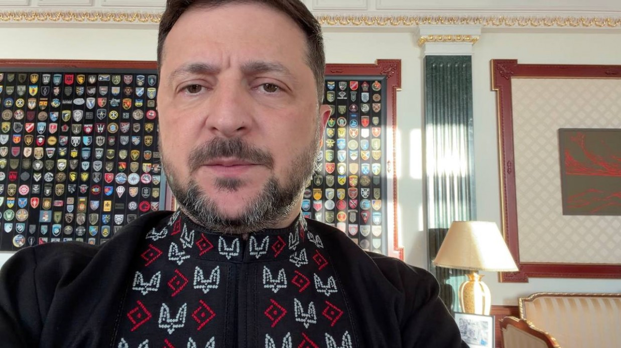 Чоловік у чорній сорочці-вишиванці з червоно-білим орнаментом зафіксований на тлі стіни з великою колекцією військових шевронів у рамках.