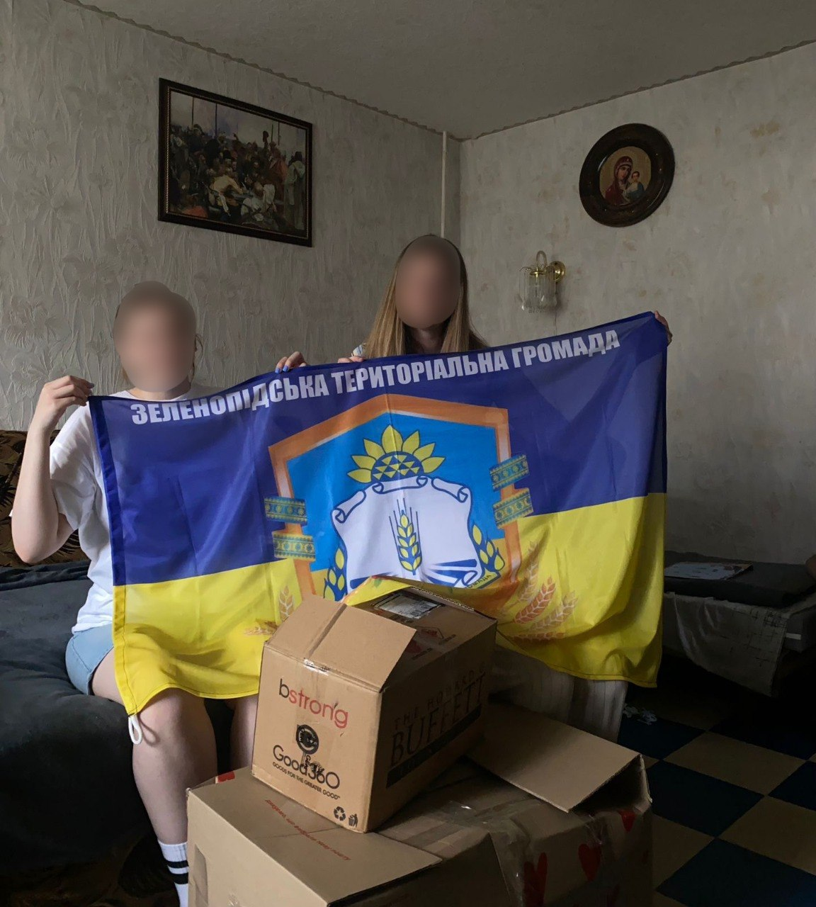 Дві людини у кімнаті тримають великий прапор Зеленопідської територіальної громади із її гербом та назвою. На передньому плані перед ними стоять картонні коробки з гуманітарною допомогою.