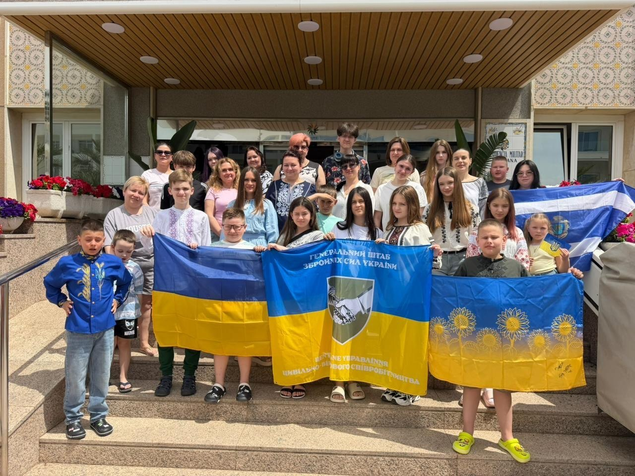 Група дорослих та дітей стоїть на сходах біля входу до будівлі, тримаючи перед собою кілька прапорів. Серед них — державний синьо-жовтий прапор України, прапор Генерального штабу ЗСУ, синій прапор із малюнком соняшників та біло-синій прапор Херсонської області.