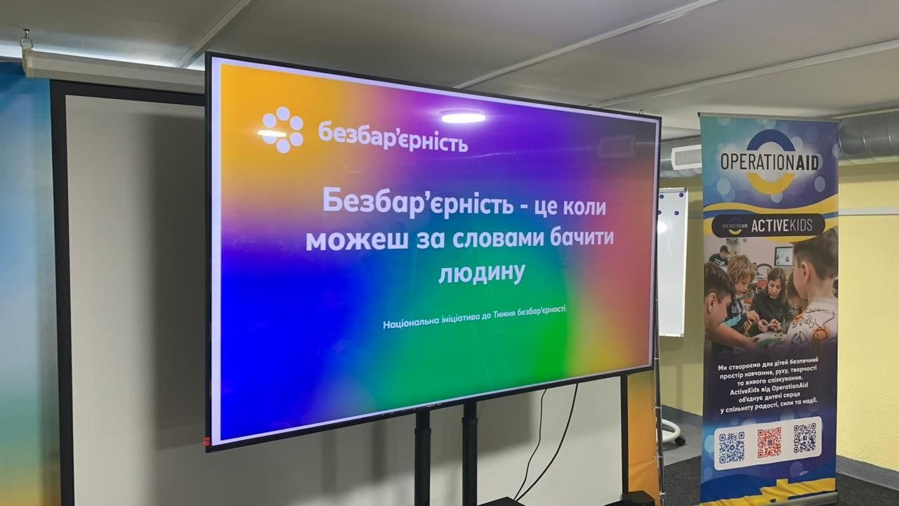 Великий яскравий екран на стійці у приміщенні демонструє слайд із гаслом «Безбар'єрність — це коли можеш за словами бачити людину». Праворуч від нього встановлено вертикальний інформаційний банер із логотипами «OPERATION AID» та «ACTIVE KIDS».