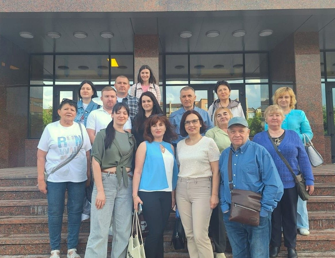 Групова фотографія людей, які стоять на гранітних сходах біля входу до будівлі з великими скляними дверима та колонами. Учасники різного віку одягнені у повсякденний літній одяг і позирують для спільного знімка.