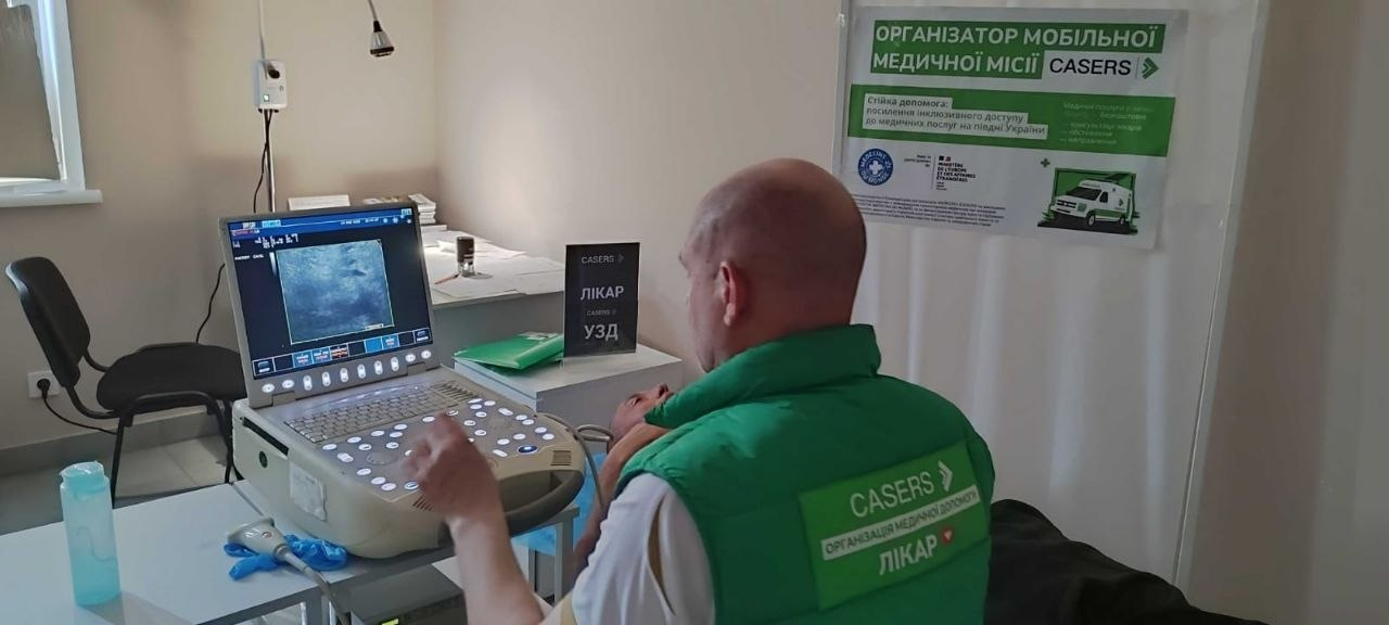 Лікар у зеленому жилеті з логотипом «CASERS» проводить ультразвукове дослідження (УЗД) пацієнту в медичному кабінеті. На стіні розміщено плакат мобільної медичної місії, а на столі — табличка з написом «Лікар УЗД».