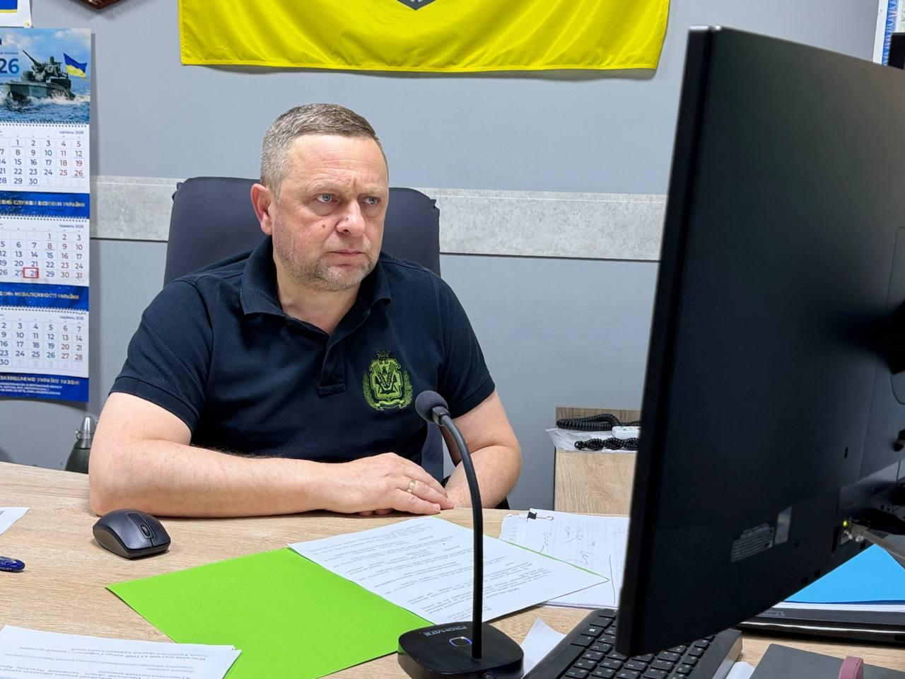Чоловік у чорній футболці-поло з шевроном сидить за робочим столом у кабінеті та дивиться на екран монітора. На столі лежать документи, комп'ютерна миша та мікрофон. На задньому плані видно настінний календар та прапор України.