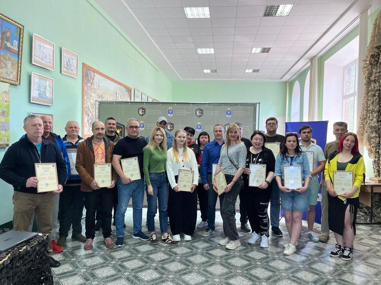 Велика група чоловіків та жінок стоїть у залі на фоні сірого та синього банерів із логотипами. Більшість присутніх тримають у руках отримані свідоцтва або грамоти у рамкаx. Вони одягнені у повсякденний одяг різного стилю та кольорів. На світло-зеленій стіні ліворуч розвішані картини.