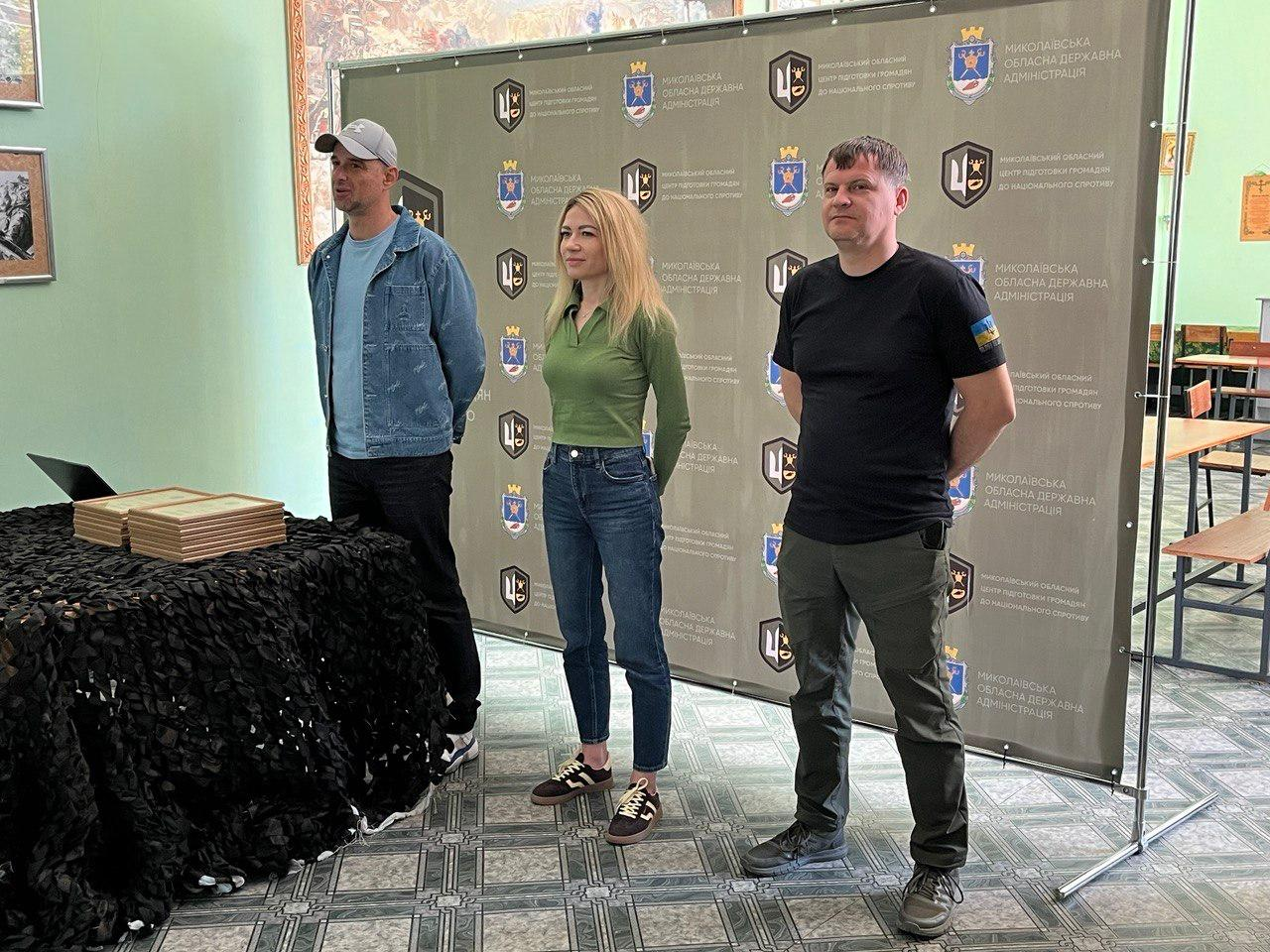 Двоє чоловіків та жінка стоять у залі на фоні великого сірого банера з логотипами. Чоловік ліворуч одягнений у джинсову куртку та кепку, жінка посередині — у зелену кофту та джинси, а чоловік праворуч — у чорну футболку та штани мілітарі. Поруч стоїть стіл із маскувальною сіткою та дипломами.