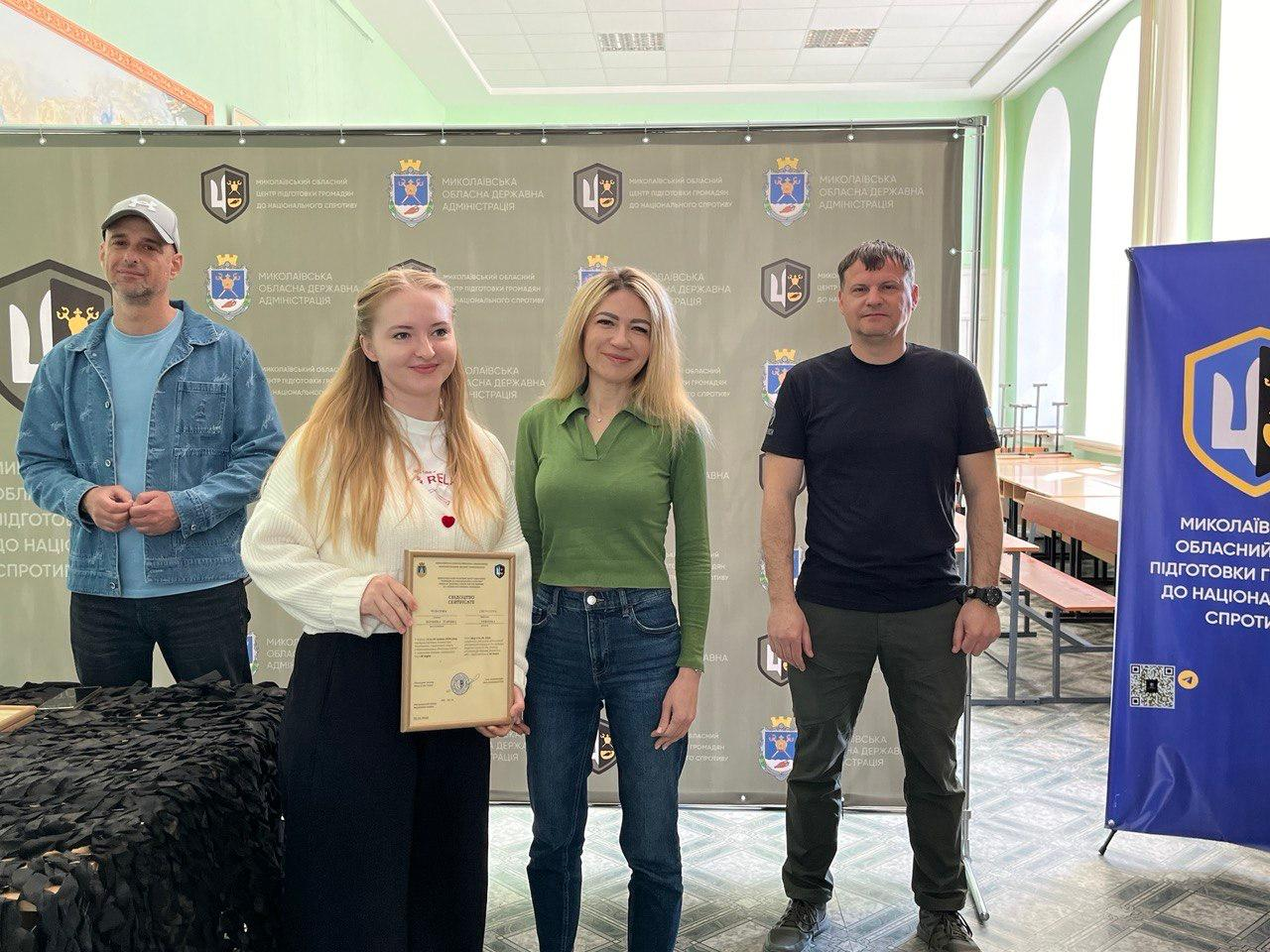 Дві жінки та двоє чоловіків стоять у залі на фоні великого сірого банера з логотипами. Молода жінка ліворуч тримає в руках свідоцтво у рамці, вона одягнена у світлий светр та темну спідницю. Поруч стоїть друга жінка у зеленій кофті з джинсами та двоє чоловіків — один у джинсовій куртці з кепкою, інший у чорній футболці та штанах мілітарі. Праворуч розташований синій вертикальний банер.