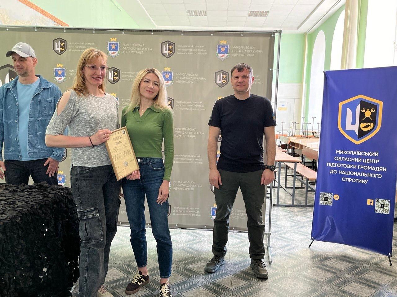 Дві жінки та двоє чоловіків стоять у залі на фоні великого сірого банера з логотипами. Жінка ліворуч тримає в руках диплом у рамці, вона одягнена у світлу кофту та темні штани. Поруч стоїть жінка у зеленій кофті з джинсами та двоє чоловіків — один у джинсовій куртці з кепкою, інший у чорній футболці та штанах мілітарі. Праворуч розташований синій вертикальний банер.