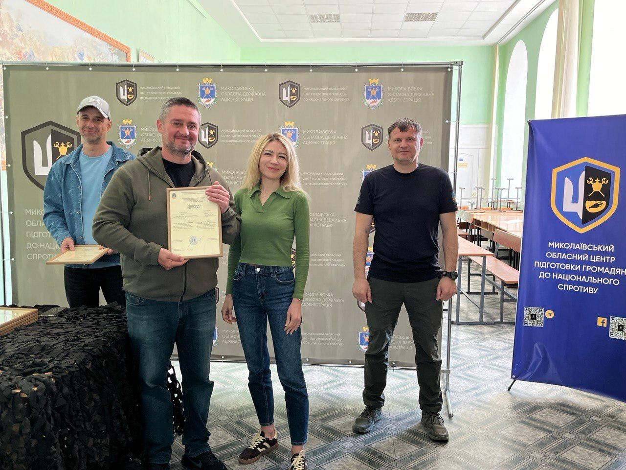 Жінка та троє чоловіків стоять у залі на фоні великого сірого банера з логотипами. Чоловік ліворуч тримає в руках свідоцтво у рамці, він одягнений у темно-зелену кофту та сині джинси. Поруч стоїть жінка у зеленій кофті з джинсами та ще двоє чоловіків — один у джинсовій куртці з кепкою, інший у чорній футболці та штанах мілітарі. Праворуч розташований синій вертикальний банер.