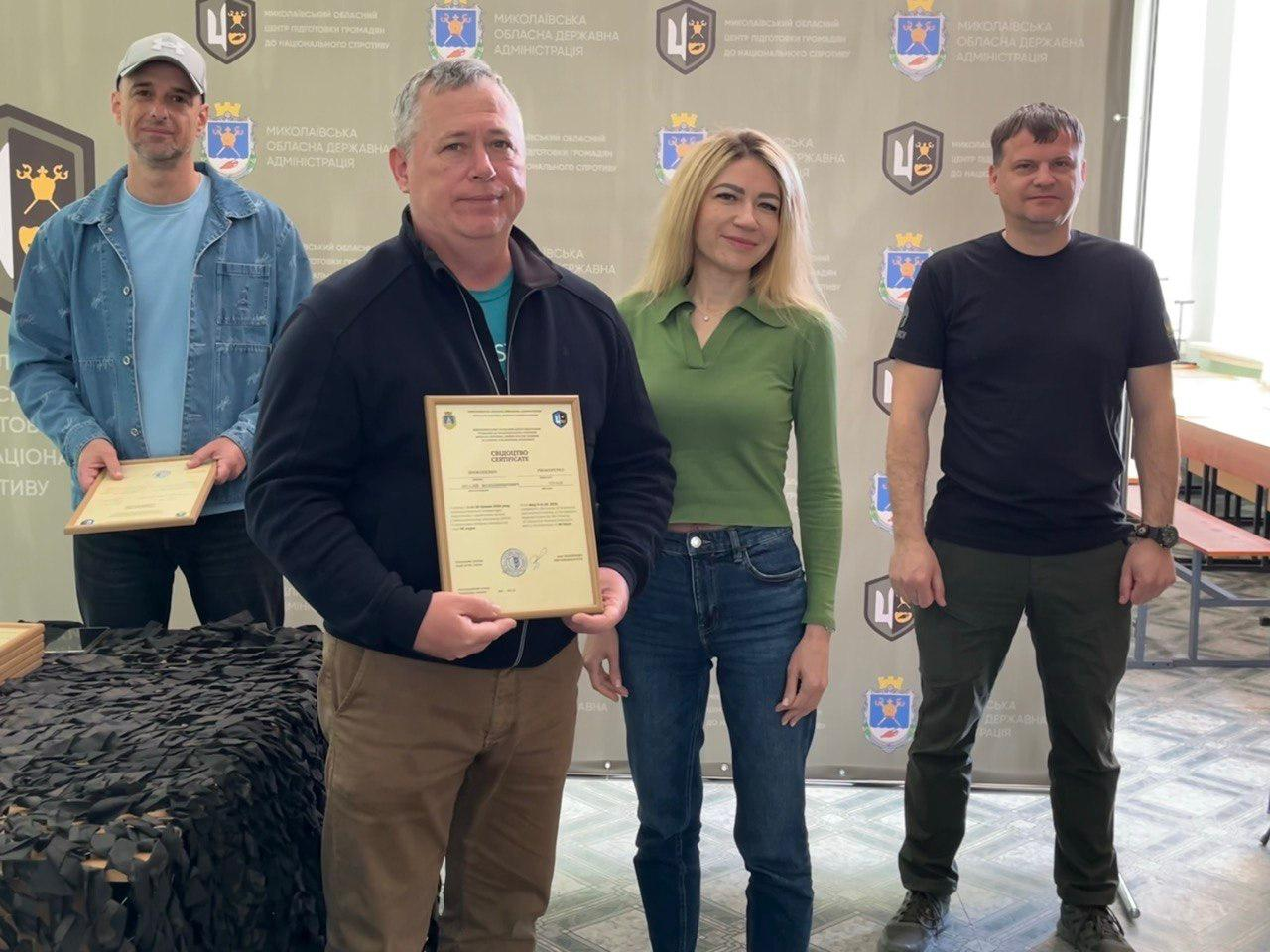 Троє чоловіків та одна жінка стоять у залі на фоні великого сірого банера з логотипами. Чоловік на передньому плані тримає в руках свідоцтво у рамці, він одягнений у чорну кофту та світлі штани. Поруч стоять жінка у зеленій кофті з джинсами та двоє чоловіків — один у джинсовій куртці з кепкою, інший у чорній футболці та штанах мілітарі.
