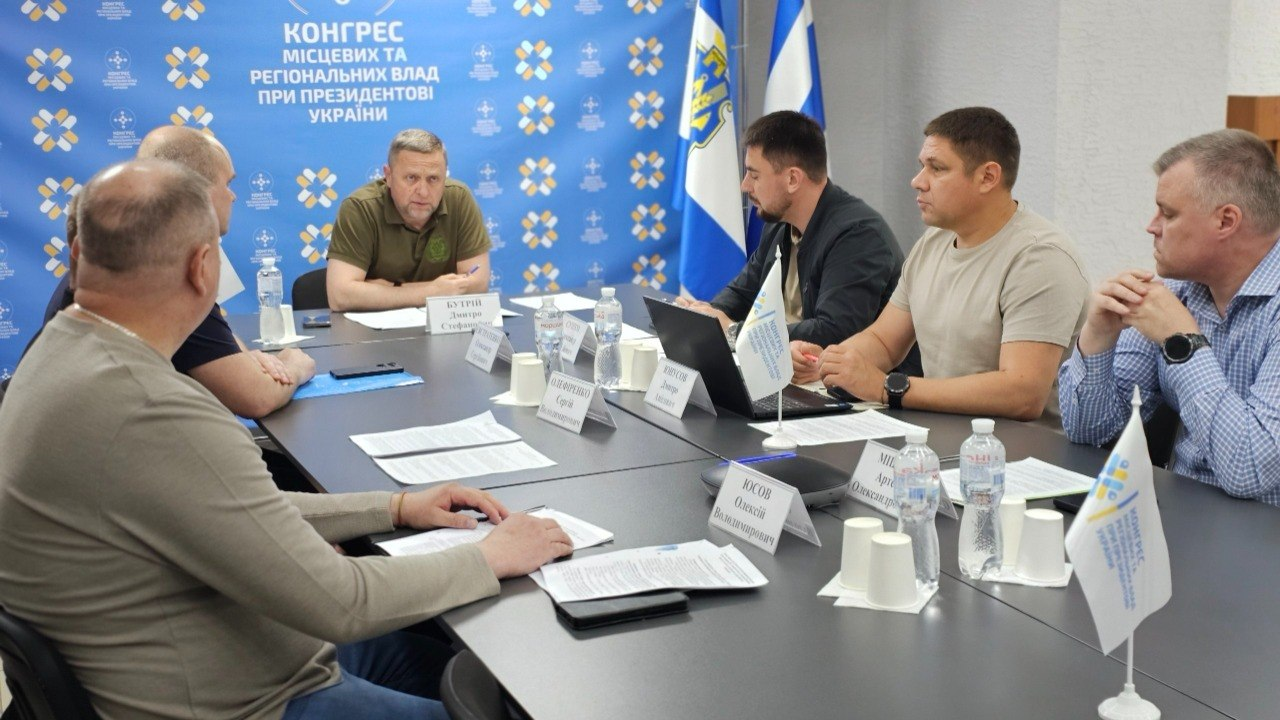 Група чоловіків сидить за Т-подібним столом під час засідання на фоні синього брендованого банера «Конгрес місцевих та регіональних влад при Президентові України» та державного прапора. На столі розміщено документи, ноутбук, пляшки з водою та паперові таблички з іменами учасників.