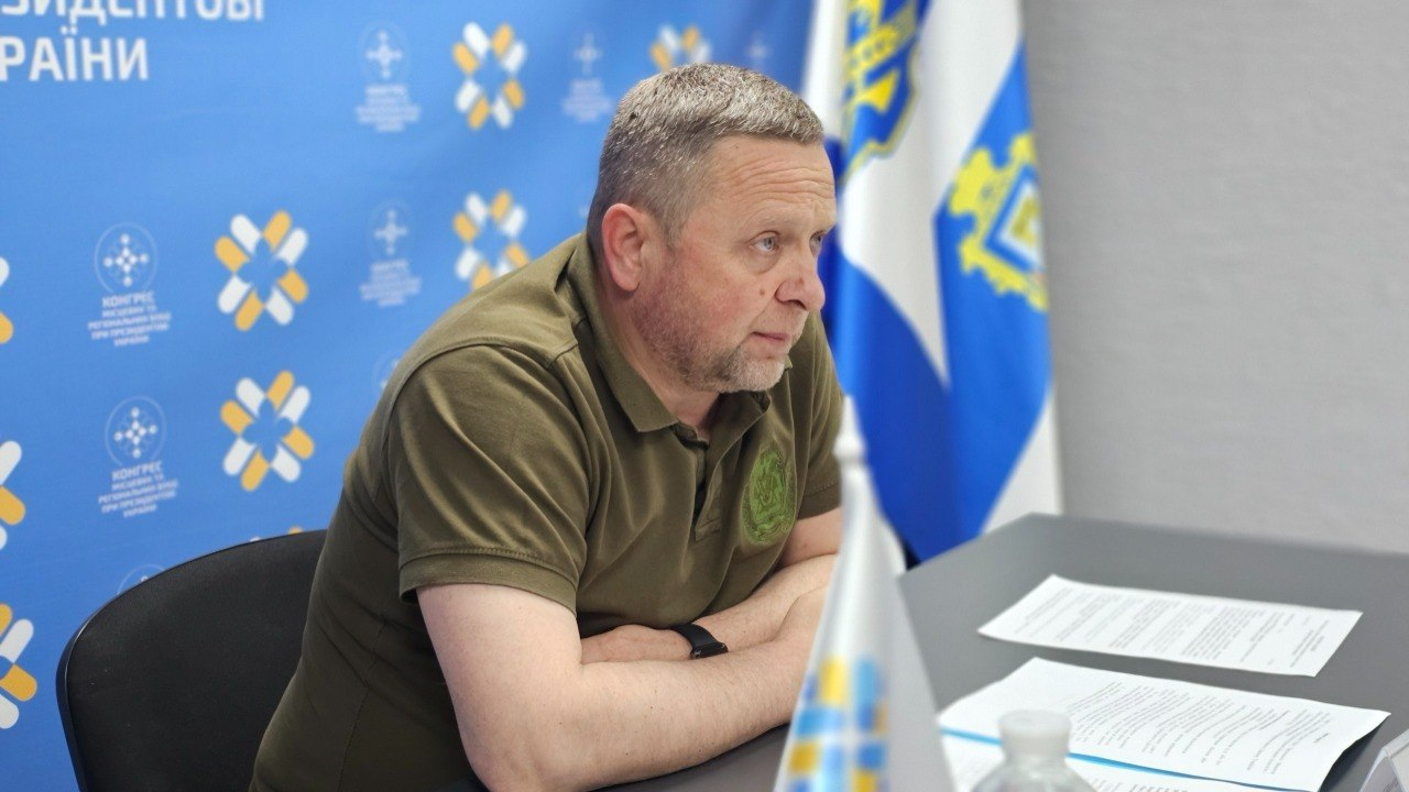 Чоловік у футболці кольору хакі сидить за столом із документами під час офіційного заходу. На задньому плані видно синій брендований банер із логотипами та прапор із гербом.