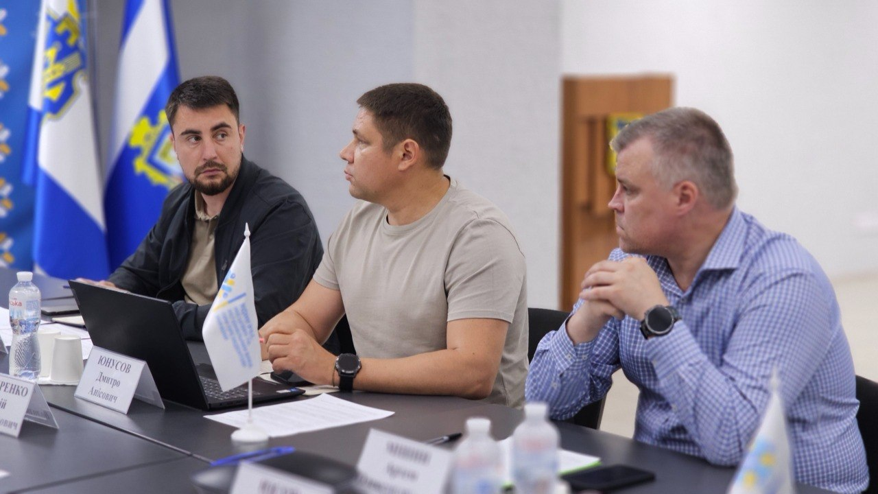 Троє чоловіків сидять поруч за робочим столом під час офіційного засідання. На столі перед ними розташовані ноутбук, документи, пляшки з водою та настільні таблички. На задньому плані видно синьо-білий прапор із гербом.