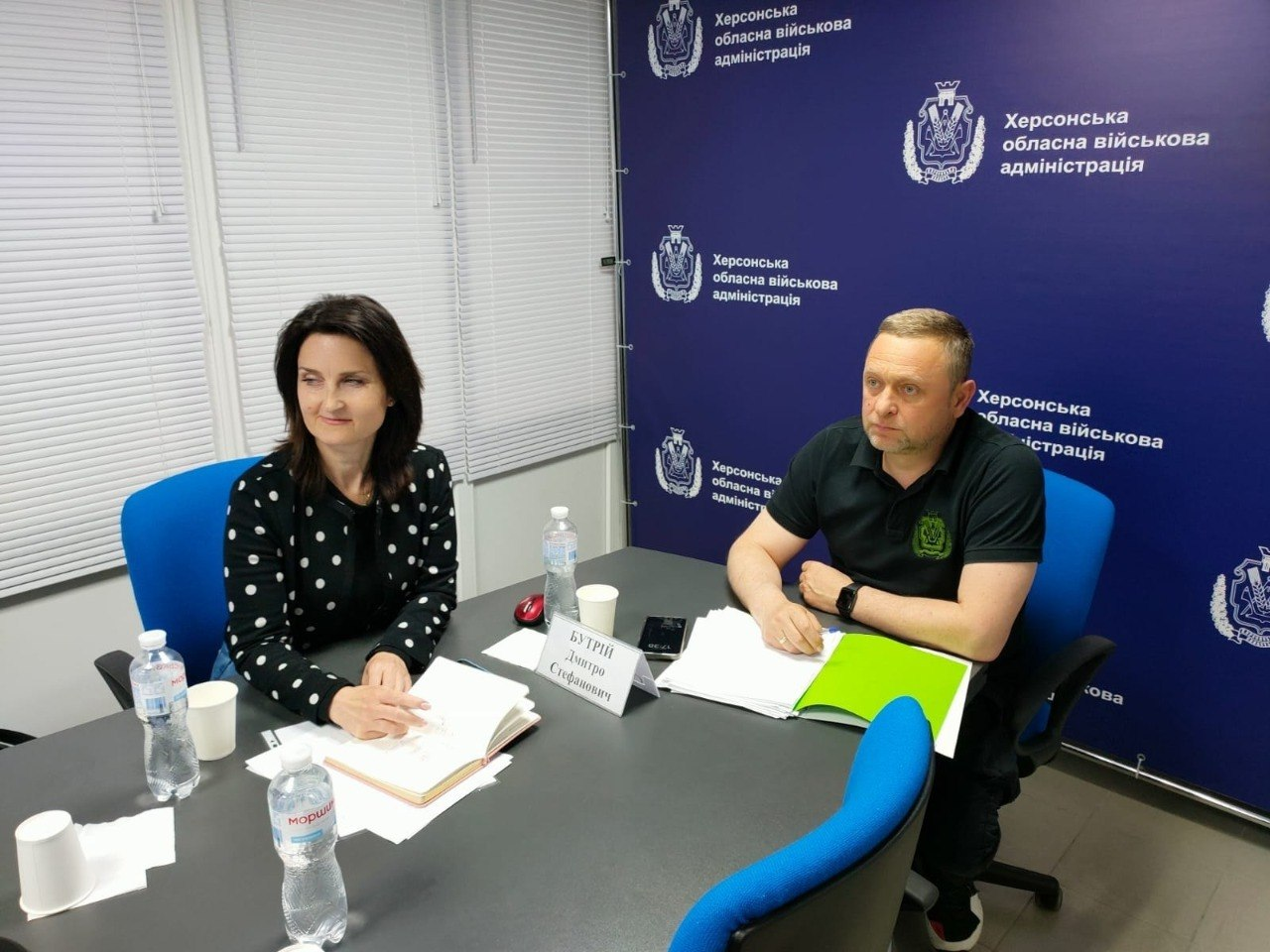 Жінка та чоловік сидять за робочим столом у кабінеті під час офіційної зустрічі чи наради. Вони слухають співрозмовника, перед ними лежать документи, папки та пляшки з водою. На задньому плані встановлено великий синій брендований банер із логотипами та офіційними написами.
