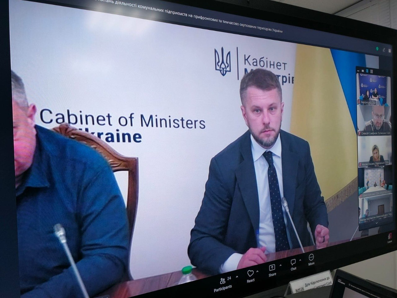 Екран великого монітора, на якому відображено онлайн-нараду у форматі відеоконференції. По центру транслюється виступ чоловіка у піджаку та білій сорочці на фоні банера Кабінету Міністрів України, ліворуч видно частину обличчя іншого учасника, а на правій бічній панелі виведені мініатюри вікон інших присутніх.