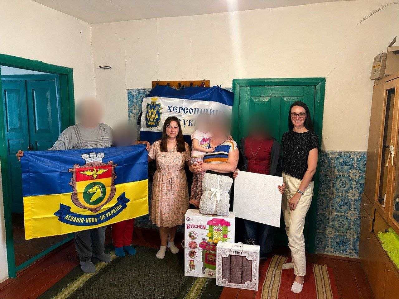 Група людей стоїть у кімнаті сільського будинку під час отримання допомоги. Обличчя чоловіка, дівчинки та жінки, яка тримає її на руках, розмиті. Дві інші жінки посміхаються в кадр. Вони тримають синьо-жовті прапори та коробки з побутовими речами й дитячою іграшковою кухнею.
