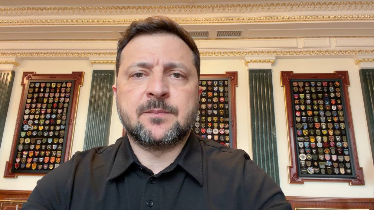 Чоловік у чорній сорочці-поло записує відеозвернення в кабінеті. На задньому плані видно класичний інтер'єр із колонами та трьома великими настінними стендами з колекцією військових шевронів.