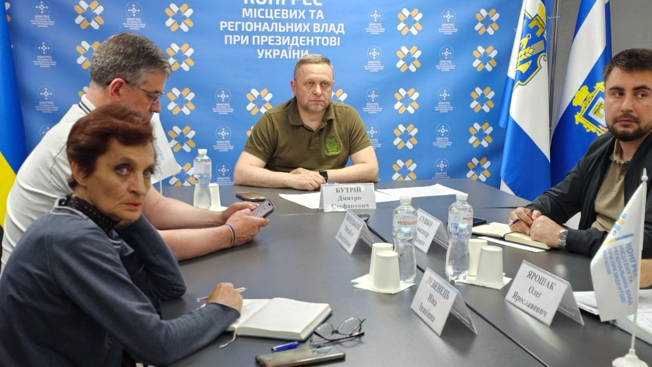 Офіційна зустріч або засідання у робочому кабінеті, де п'ятеро людей сидять навколо великого столу з документами, пляшками води та блокнотами. На задньому плані розміщено синій банер із логотипами та офіційним текстом, а також державну символіку.