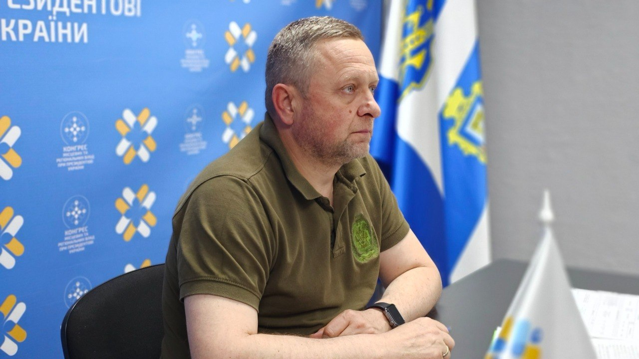 Чоловік середнього віку у футболці кольору хакі сидить за столом, склавши руки перед собою, та дивиться вбік. На задньому плані ліворуч розміщено синій стенд із логотипами, а праворуч — синьо-жовтий прапор із гербом.