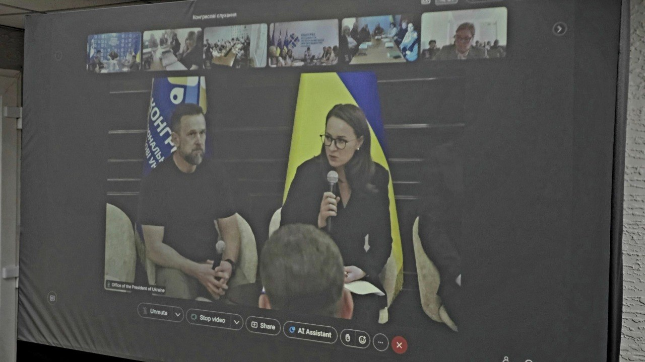 Проєкція онлайн-трансляції на великому екрані в приміщенні, де відображається відеоконференція за участю спікерів та інших підключених учасників. У центрі кадру жінка в окулярах говорить у мікрофон, поруч із нею сидить чоловік, а на задньому плані видно прапори.