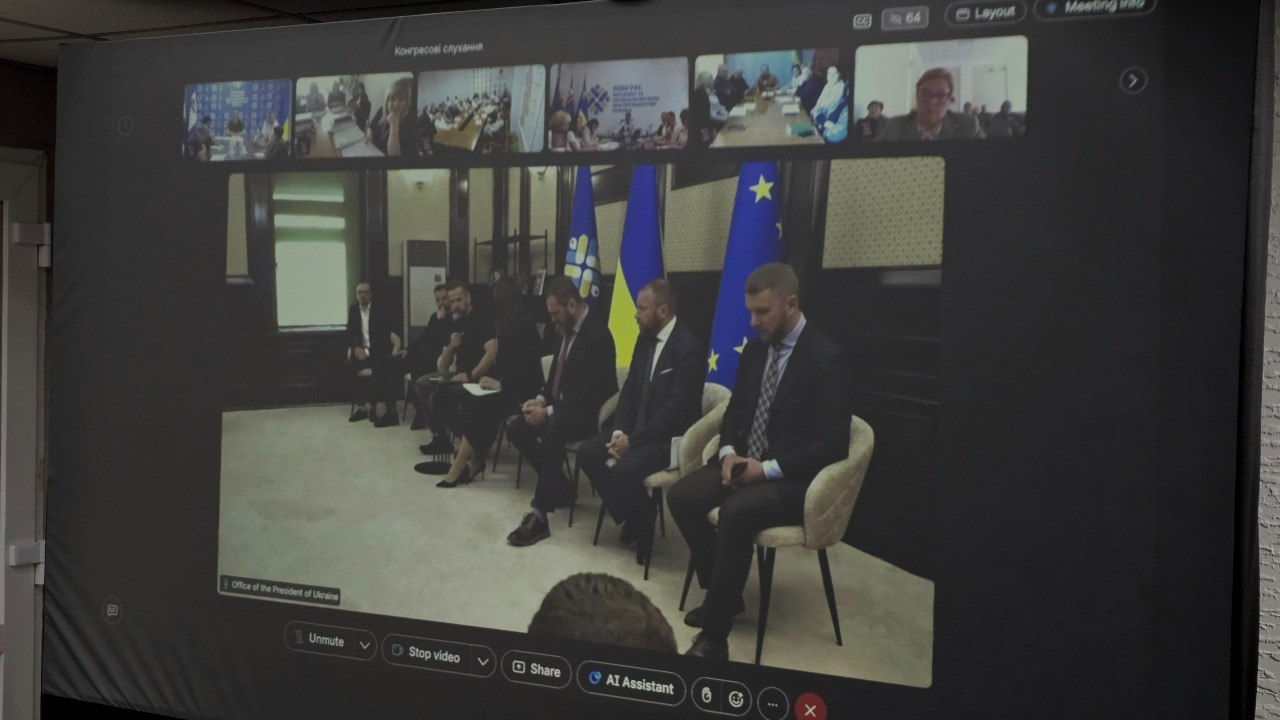 Проєкція онлайн-трансляції на великому екрані, де відображається офіційна зустріч. У центрі кадру видно групу чоловіків у костюмах, які сидять на стільцях перед прапорами, а зверху та знизу розміщено елементи інтерфейсу відеоконференції.