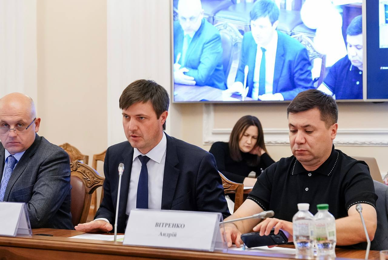 Офіційне засідання, де троє чоловіків сидять за столом із мікрофонами, табличками та пляшками води. На задньому плані видно великий екран із трансляцією відеодзвінка та жінку, яка робить записи.