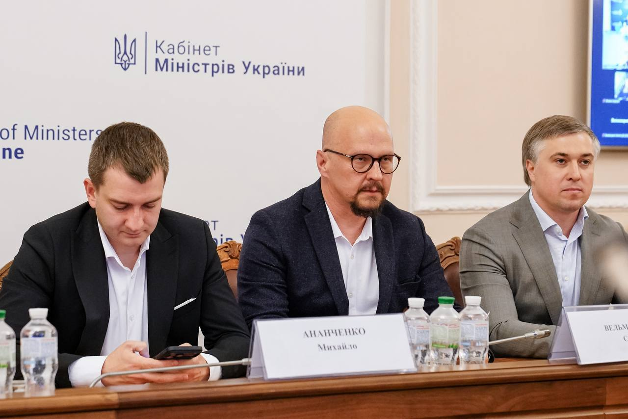 Троє чоловіків у піджаках сидять за столом під час засідання на тлі стіни з написом «Кабінет Міністрів України». На столі перед ними встановлено мікрофони, пляшки з водою та настільні таблички.