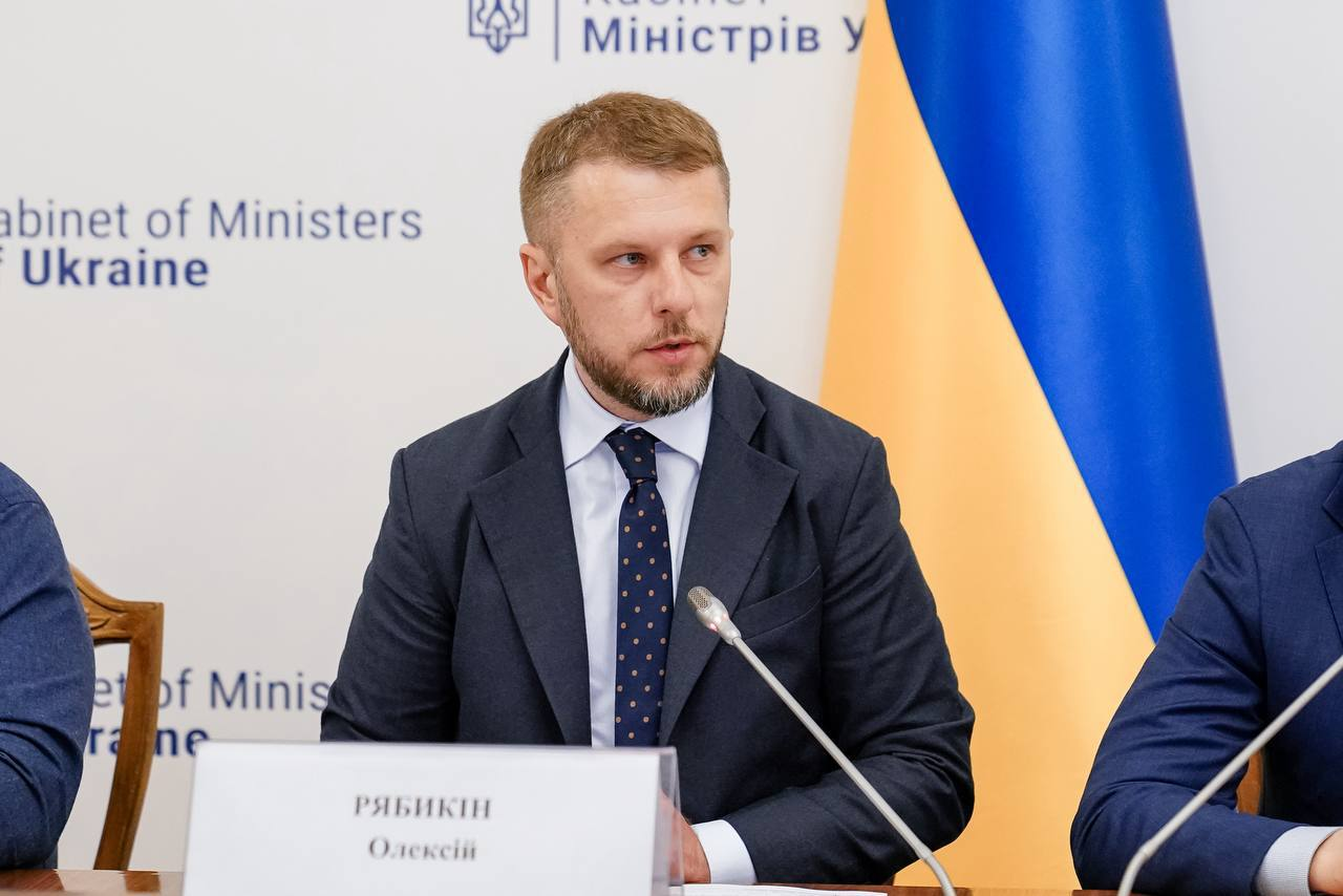 Чоловік у темному костюмі, білій сорочці та краватці виступає перед мікрофоном за столом засідань. На білому фоні позаду нього видно частину державного прапора України та офіційний напис «Cabinet of Ministers of Ukraine».