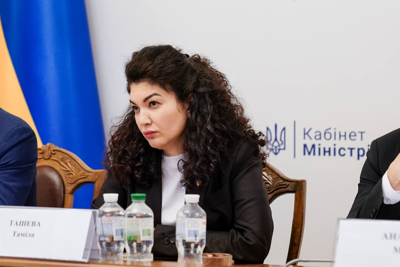 Жінка з кучерявим волоссям у темному піджаку сидить за столом президії під час офіційного заходу. На фоні видно Державний Прапор України та напис «Кабінет Міністрів».