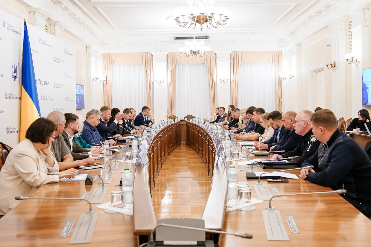 Офіційне засідання у світлій залі, де учасники сидять по обидва боки великого U-подібного дерев'яного столу з мікрофонами, документами та пляшками води. На лівому фоні розміщено синій Державний Прапор України та банер із гербом.