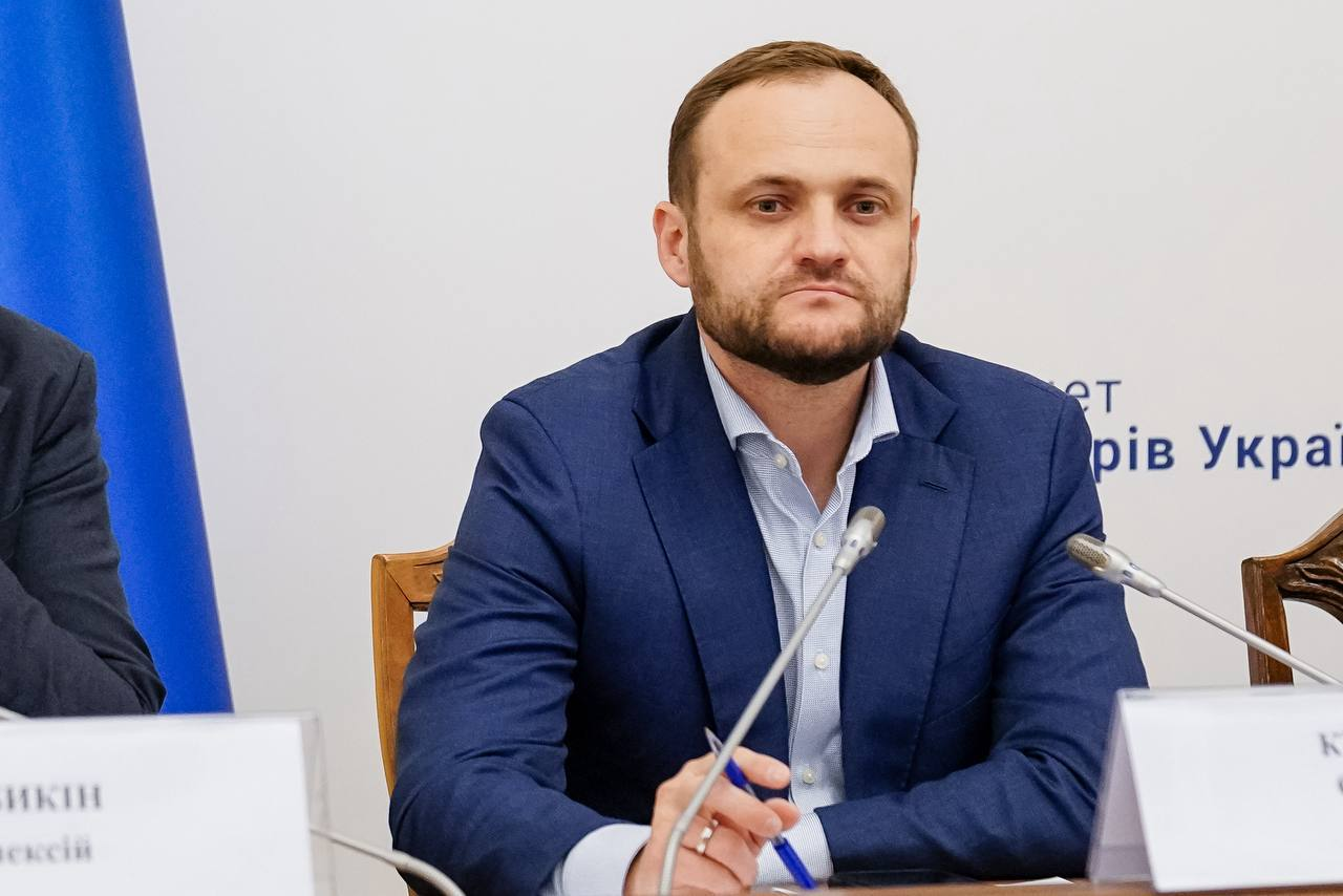Чоловік у синьому класичному костюмі та сорочці сидить за столом президії перед мікрофонами, тримаючи в руці ручку. На задньому плані частково видно офіційний напис українською мовою та синій елемент декору.