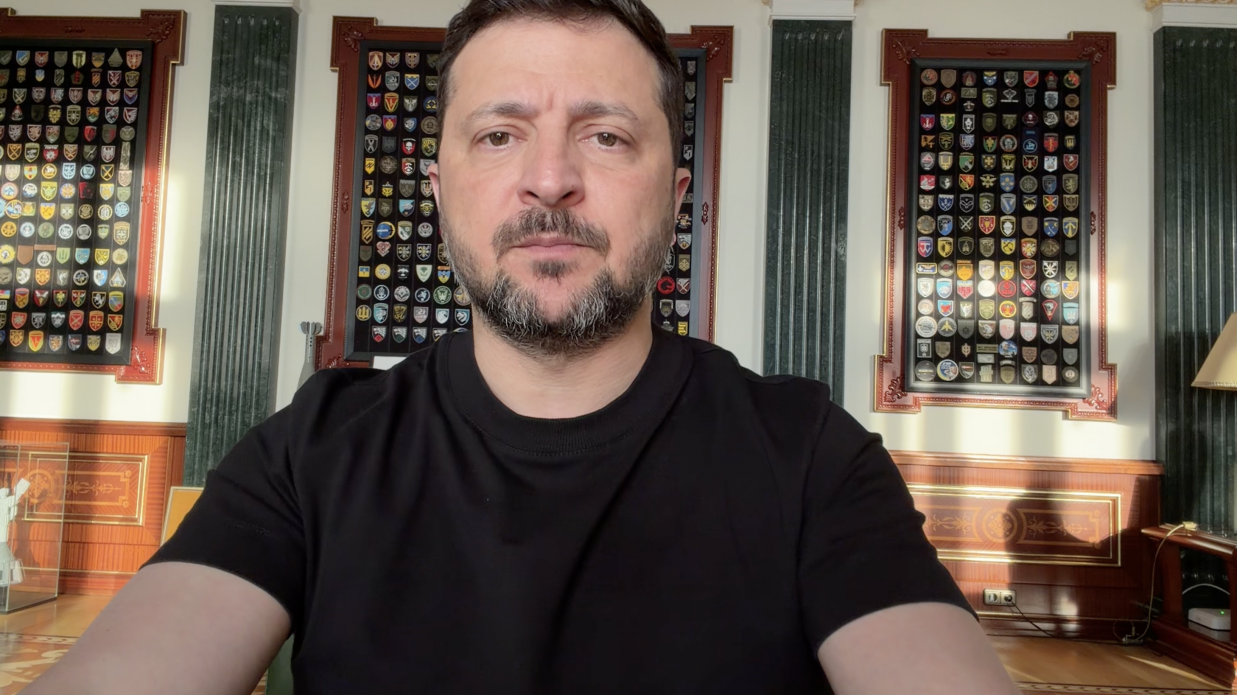 Чоловік із короткою бородою та в чорній футболці дивиться прямо в камеру. Він перебуває в кабінеті з класичним дерев'яним інтер'єром та зеленими колонами. На стіні за ним розміщені три великі вертикальні рамки, заповнені численними різнокольоровими військовими шевронами та нашивками.