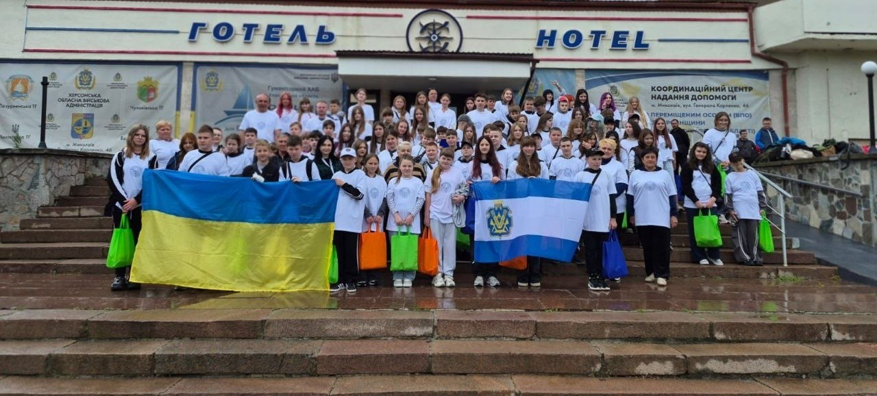 Велика група дітей та дорослих у білих футболках стоїть на широких кам'яних сходах перед будівлею з написом «ГОТЕЛЬ / HOTEL», тримаючи в руках розгорнуті державний прапор України та прапор Херсонської області, а також яскраві кольорові пакети на тлі інформаційних банерів координаційного центру допомоги.