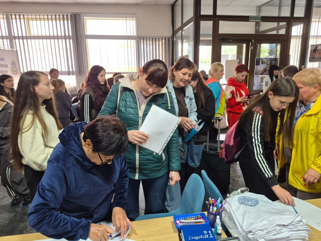 Група людей у просторому світлому приміщенні біля столу реєстрації, де жінка на передньому плані заповнює документи, а інші відвідувачі переглядають папери та чекають у черзі поруч із розкладеними речами.