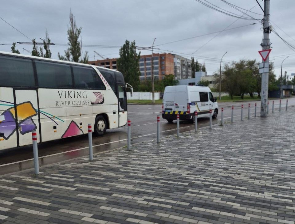 Великий білий туристичний автобус із написом «VIKING RIVER CRUISES» та білий поліцейський мікроавтобус стоять на мокрому асфальті біля викладеного бруківкою тротуару з обмежувальними стовпчиками в похмуру погоду.