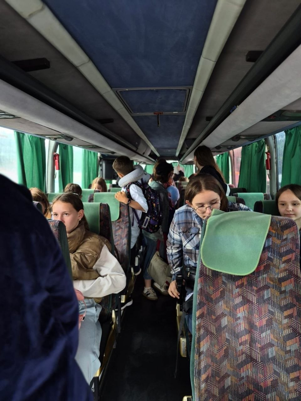 Група дітей та підлітків із рюкзаками розміщується в салоні пасажирського автобуса із зеленими шторами та візерунчастими сидіннями.