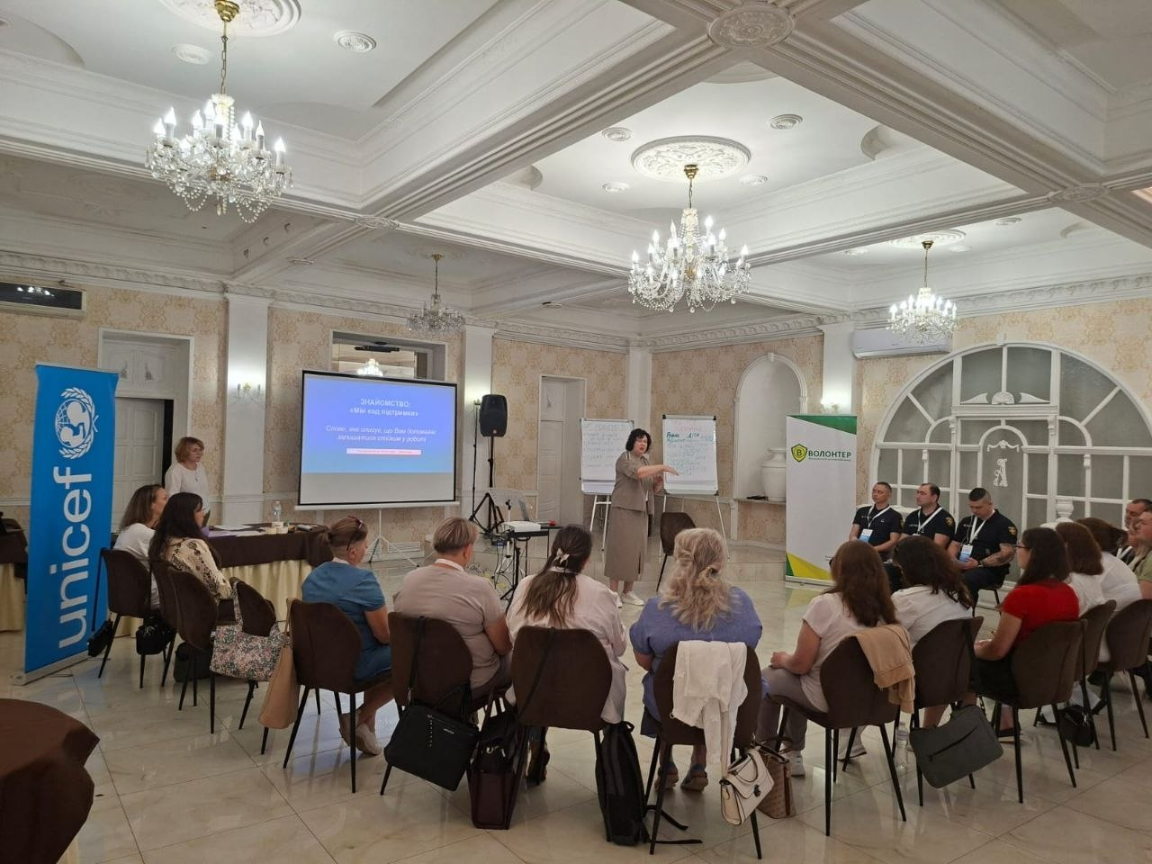 Урочиста зала з кришталевими люстрами, де проходить тренінг або презентація. Група людей сидить півколом на стільцях спіною до камери, слухаючи жінку-спікера, яка стоїть попереду біля проєкційного екрана та фліпчартів. Зліва встановлено синій вертикальний банер ЮНІСЕФ (Unicef).