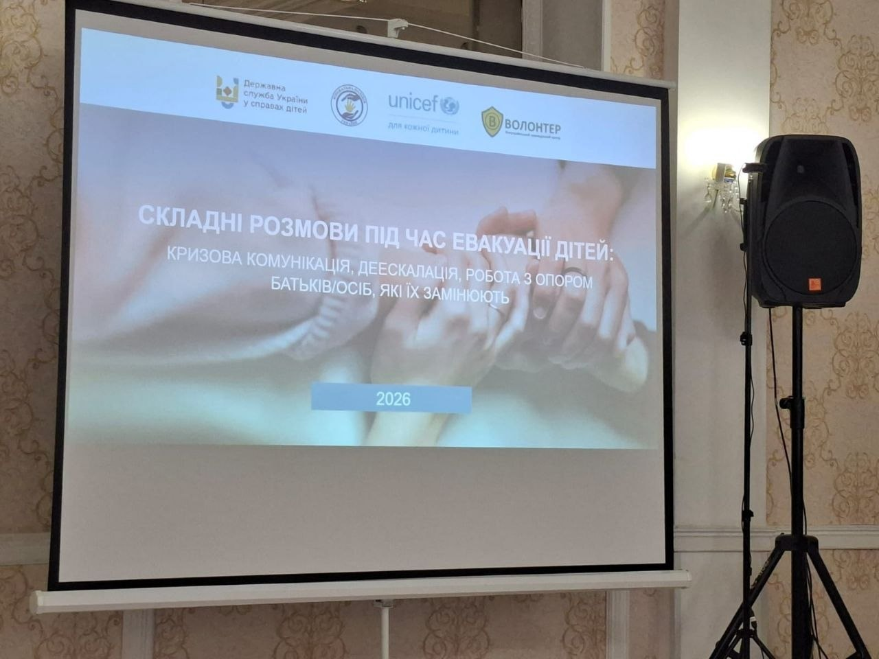 Проєкційний екран у залі з титульним слайдом презентації на тему: «Складні розмови під час евакуації дітей: кризова комунікація, деескалація, робота з опором батьків/осіб, які їх замінюють». Зверху розміщено логотипи партнерів (зокрема ЮНІСЕФ), а праворуч стоїть акустична колонка.