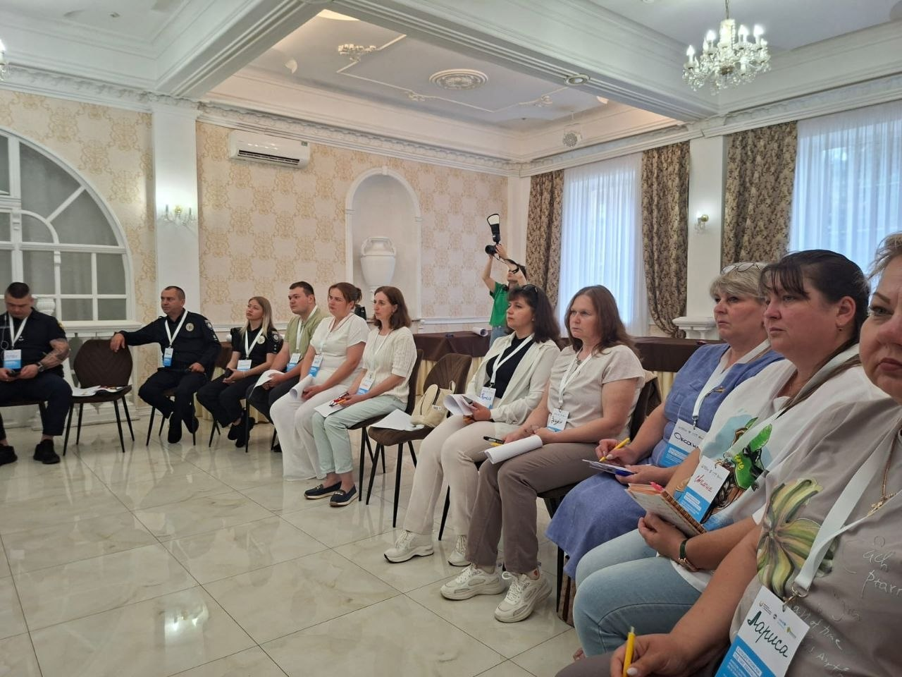 Група учасників офіційного заходу сидить на стільцях уздовж стіни у світлій залі з класичним інтер'єром. Серед присутніх — цивільні особи та працівники поліції у формі, які слухають презентацію або спікера. На передньому плані видно людей із блокнотами, ручками та бейджами.