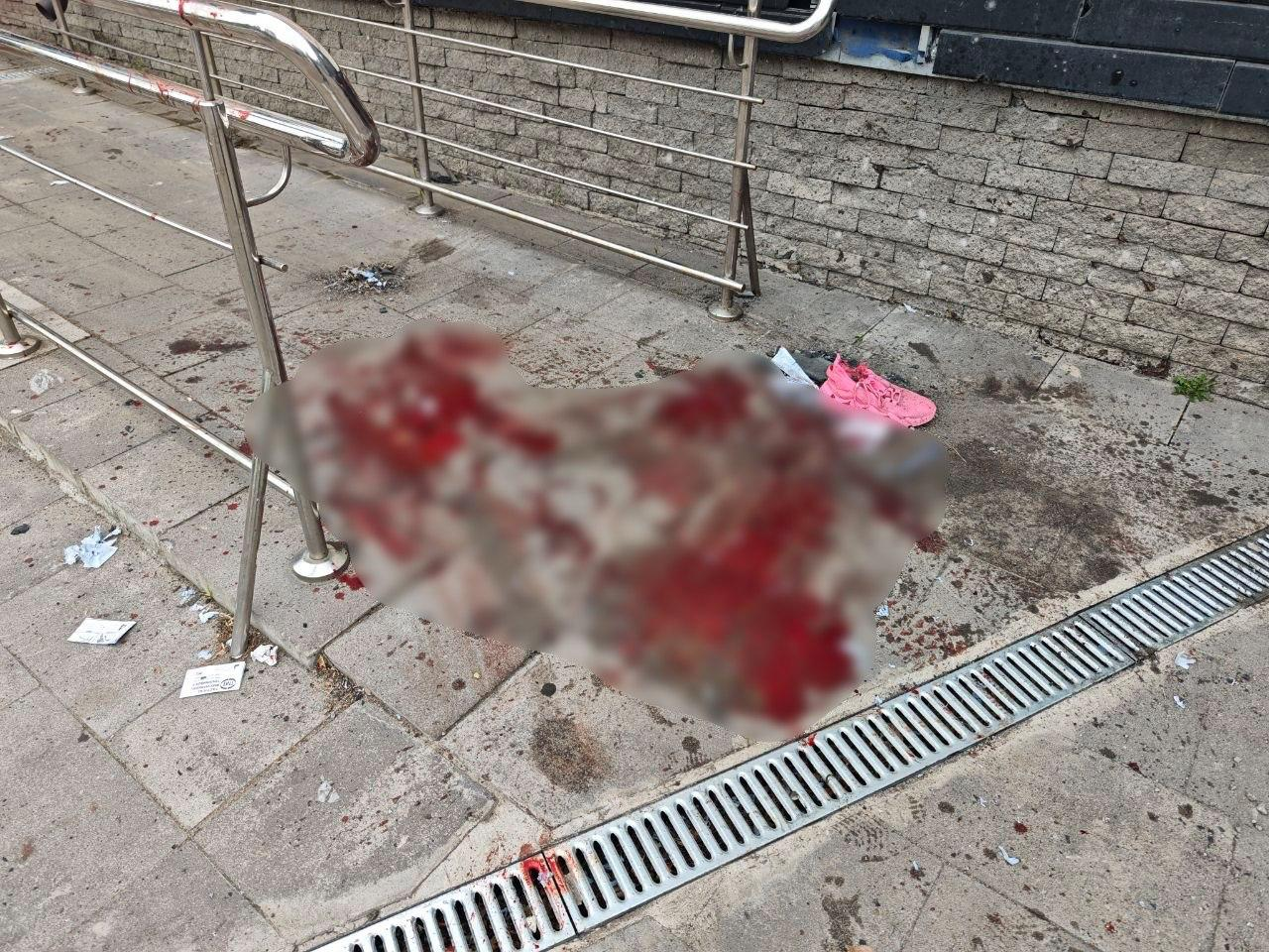 Місце події на вулиці біля металевого огородження-пандуса та кам'яної стіни. На плитці лежить великий розмитий об'єкт із червоними плямами, навколо якого видно численні сліди крові, дрібне сміття та один рожевий кросівок.