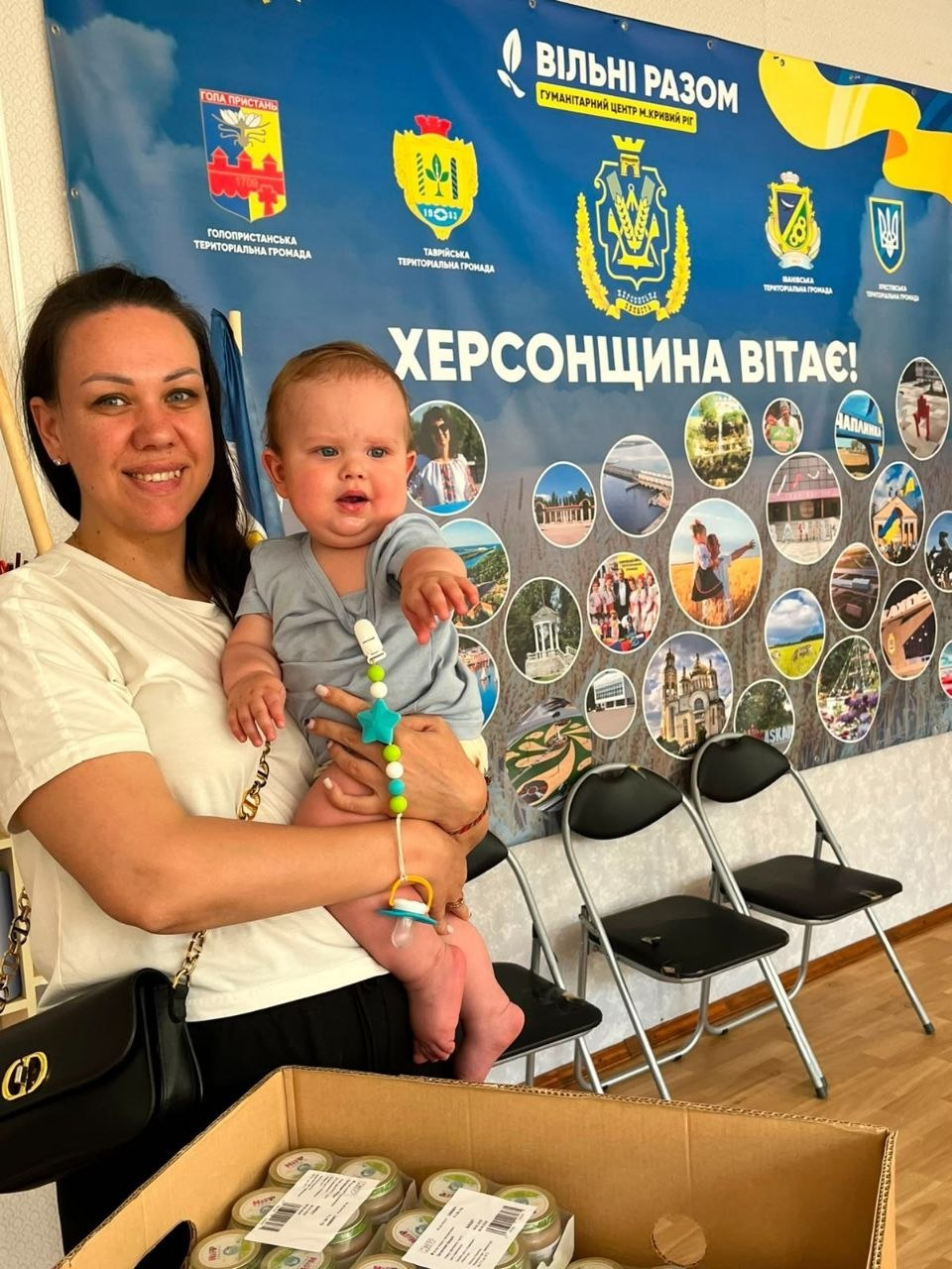 Жінка з немовлям на руках стоїть на тлі великого синьо-жовтого банера «Херсонщина вітає!» у гуманітарному центрі. На передньому плані біля них стоїть відкрита картонна коробка з баночками дитячого харчування.