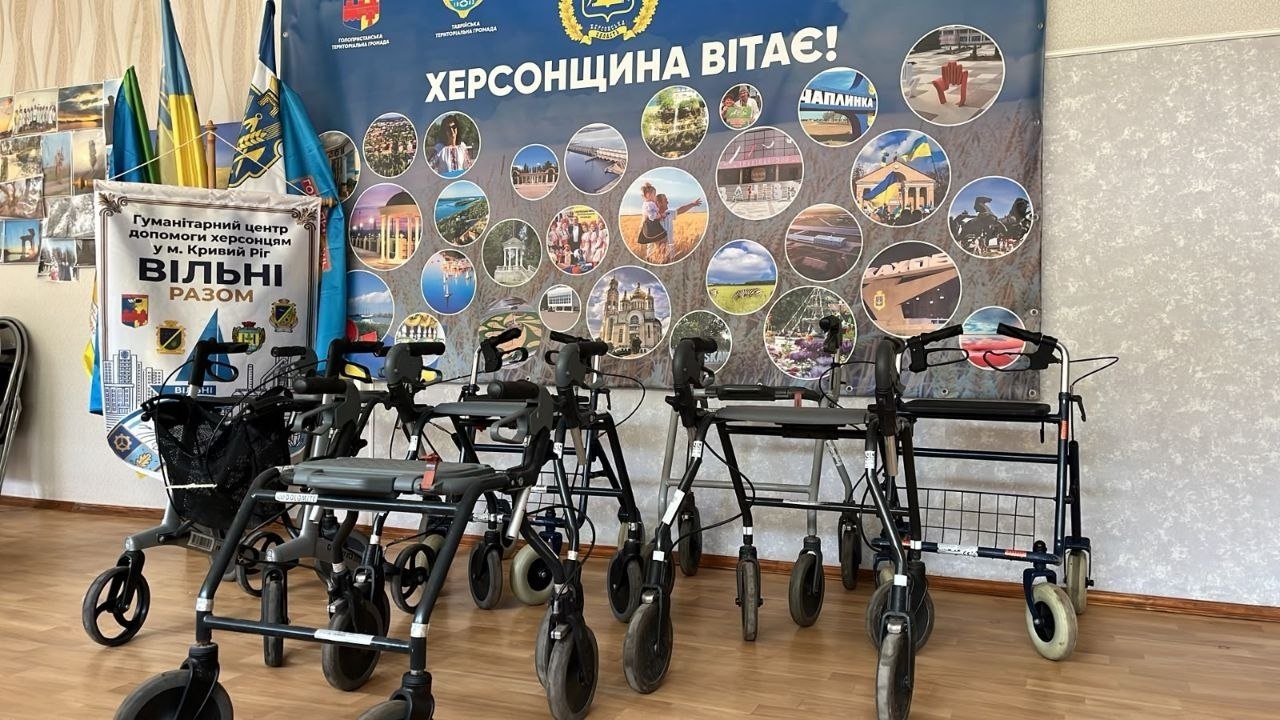 Ряд чотириколісних ходунків-ролаторів виставлено у приміщенні гуманітарного центру «Вільні разом». На задньому плані видно державні прапори та великий синій банер з написом «Херсонщина вітає!».