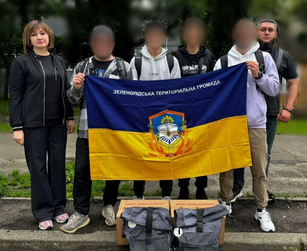 Група людей стоїть на вулиці та тримає синьо-жовтий прапор із написом «Зеленопідська територіальна громада» та її гербом. Попереду на асфальті біля їхніх ніг стоять картонні коробки з написом «Продуктовий набір» та два сірі рюкзаки.
