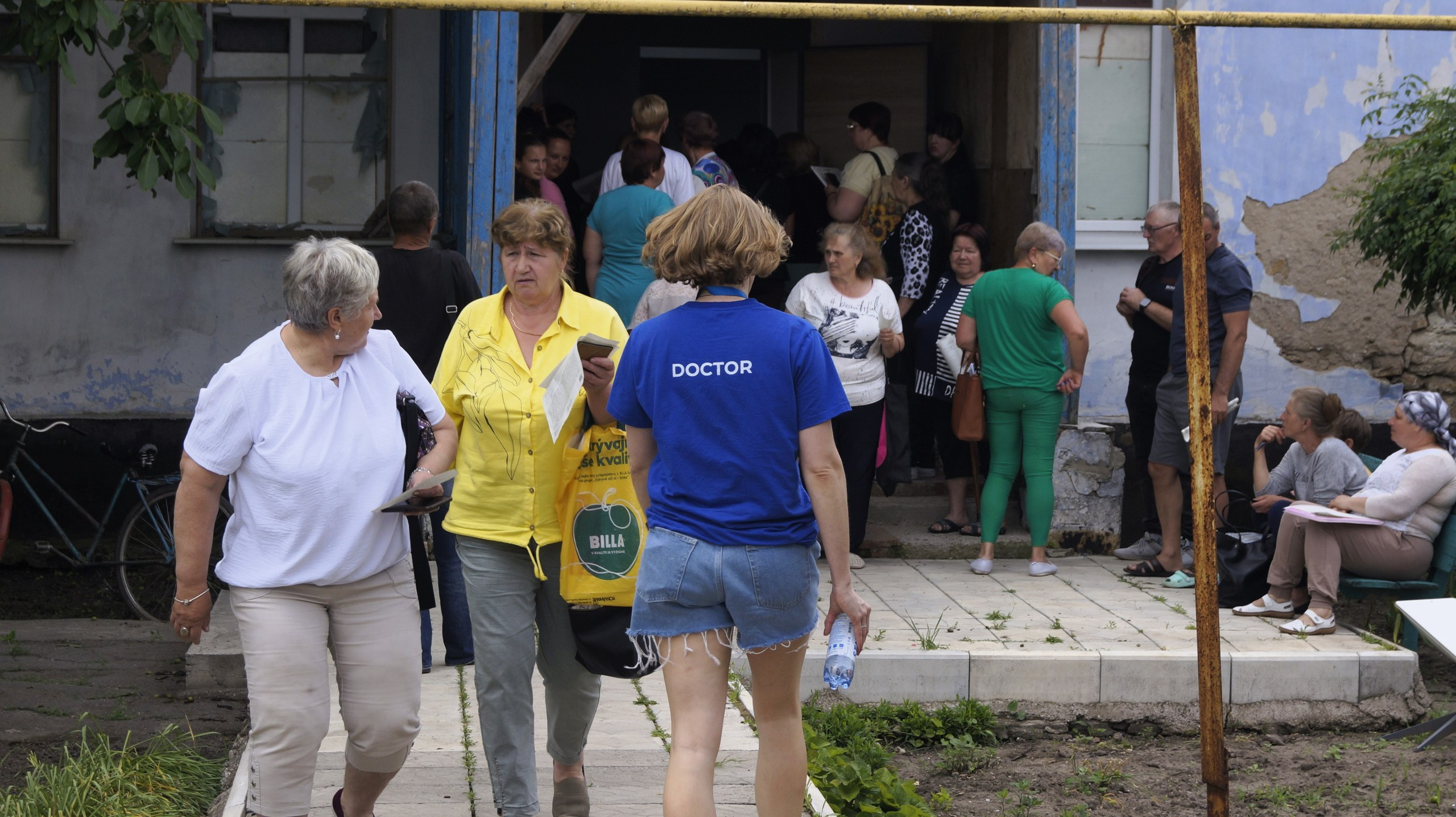 Група людей різного віку біля входу до будівлі, перед якими спиною до камери йде медичний працівник у синій футболці з написом «DOCTOR».