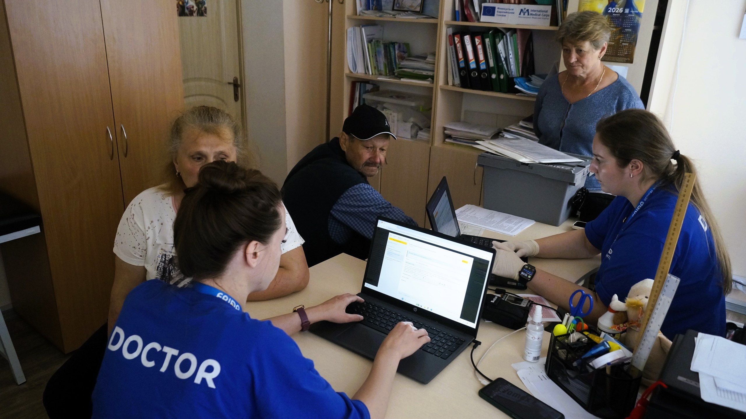 Медичні працівники у синій уніформі з написом «DOCTOR» ведуть прийом громадян похилого віку за столом з ноутбуками в офісному приміщенні.