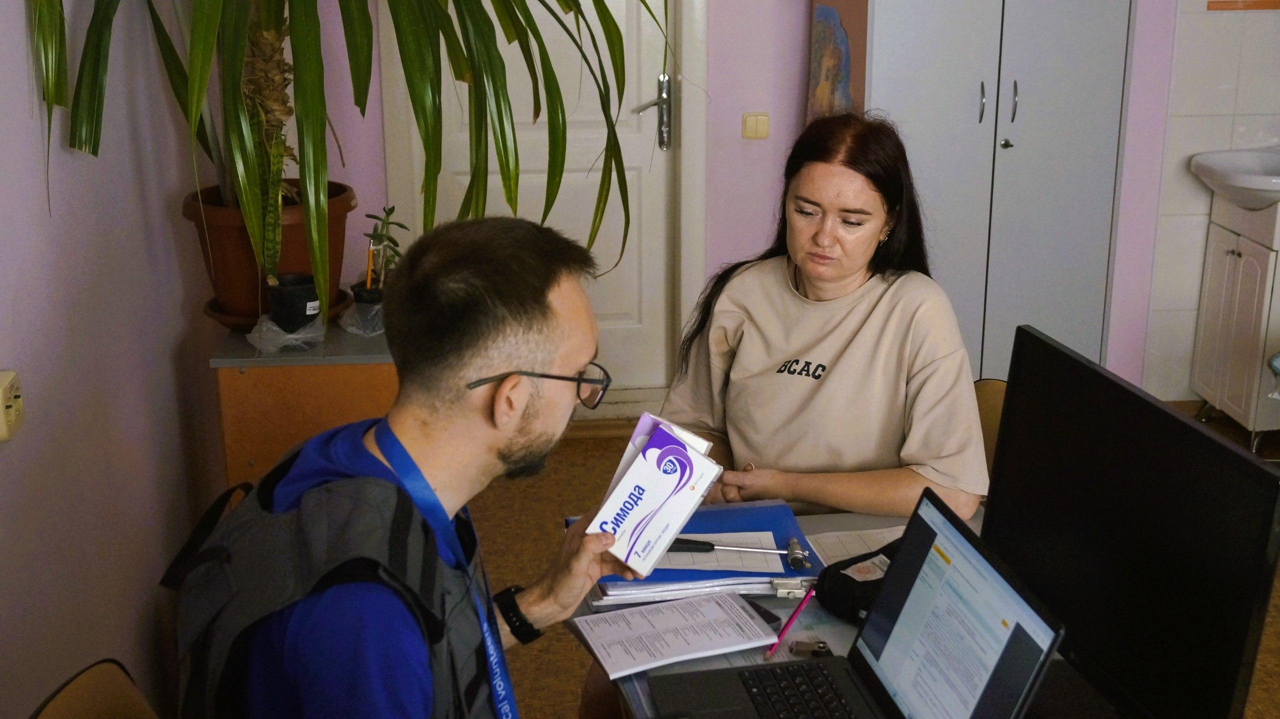 Чоловік у синій формі медичного волонтера консультує жінку за столом, тримаючи в руці упаковку лікарського засобу «Симода».