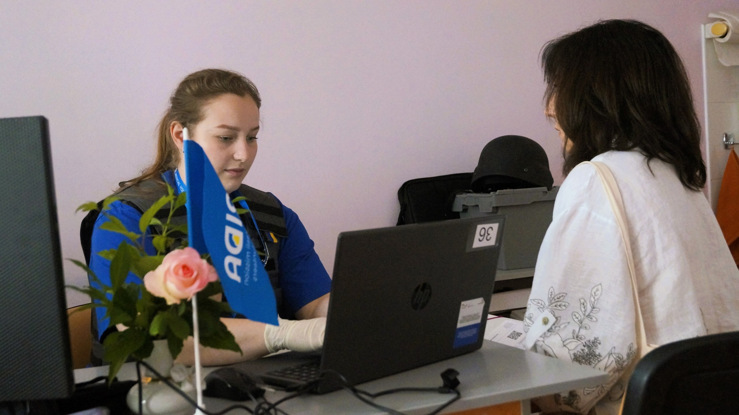 Жінка-медик у синій формі волонтера організації «FRIDA» консультує пацієнтку за робочим столом із ноутбуком, на якому встановлено маленький синій прапорець.