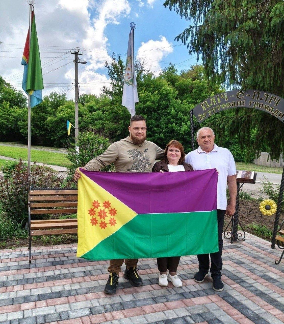 Три людини тримають прапор із фіолетовою, зеленою та жовтою смугами і червоними зірками на тлі металевої арки з написом «ПЛІЧ-О-ПЛІЧ згуртовані громади» та двома прапорами на флагштоках.